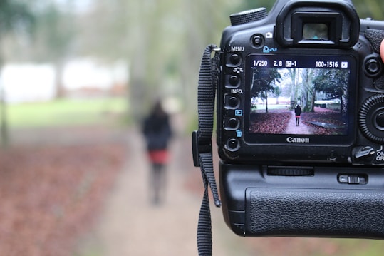 A digital camera is positioned in the foreground with the viewfinder screen displaying an image of a person standing on a path in a park. The background outside the camera display is blurred, featuring the same path and surrounding greenery.