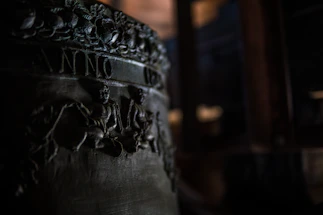A close-up view of an engraved metal surface, possibly a part of a bell with detailed relief carvings. The image shows intricate designs of figures and foliage, with the word 'ANNO' partially visible. The lighting is dim, highlighting the textures and engravings.