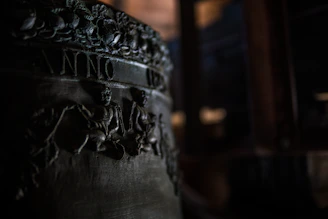 A close-up view of an engraved metal surface, possibly a part of a bell with detailed relief carvings. The image shows intricate designs of figures and foliage, with the word 'ANNO' partially visible. The lighting is dim, highlighting the textures and engravings.