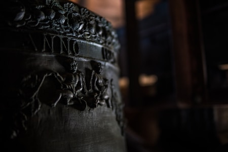 A close-up view of an engraved metal surface, possibly a part of a bell with detailed relief carvings. The image shows intricate designs of figures and foliage, with the word 'ANNO' partially visible. The lighting is dim, highlighting the textures and engravings.