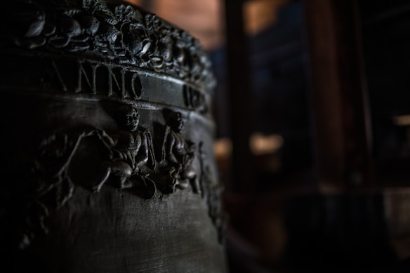 A close-up view of an engraved metal surface, possibly a part of a bell with detailed relief carvings. The image shows intricate designs of figures and foliage, with the word 'ANNO' partially visible. The lighting is dim, highlighting the textures and engravings.