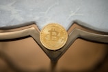A golden Bitcoin token is placed against a reflective surface with geometric shapes and a lightly textured background.