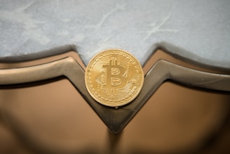 A golden Bitcoin token is placed against a reflective surface with geometric shapes and a lightly textured background.