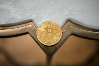 A golden Bitcoin token is placed against a reflective surface with geometric shapes and a lightly textured background.