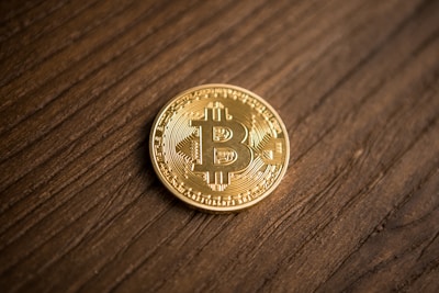 A single gold-colored coin featuring the Bitcoin symbol rests on a wooden surface with a natural, grainy texture. The coin has detailed engravings and is prominently displayed against the simple wooden background.