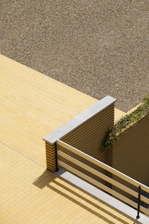 A top-down view of a minimalist outdoor space featuring a yellow tiled floor, a beige gravel area, and a section of a brick wall with horizontal stripes. The area includes a portion of a railing and a small vertical section of the wall covered with greenery.