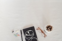 A flatlay of office supplies arranged on a light surface. Items include a black notebook with white earphones, a pair of tortoiseshell glasses, a white container holding colorful pencils, a pen, and scattered paper clips in various colors along with a binder clip and a sticky note.