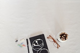 A flatlay of office supplies arranged on a light surface. Items include a black notebook with white earphones, a pair of tortoiseshell glasses, a white container holding colorful pencils, a pen, and scattered paper clips in various colors along with a binder clip and a sticky note.