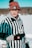 A person wearing a striped referee shirt, gray hoodie, and a red knit hat stands outside in a snowy environment. They are holding a can of Coors Light in one hand and wearing black gloves with a logo. The background appears to be a snowy landscape with trees.