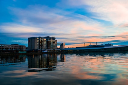 A calm waterfront scene with a modern hotel building situated along a riverbank. The building reflects in the still water, under a sky painted with soft pastel hues of pink, orange, and blue during sunset.