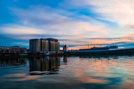 A calm waterfront scene with a modern hotel building situated along a riverbank. The building reflects in the still water, under a sky painted with soft pastel hues of pink, orange, and blue during sunset.