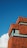 A modern building with a distinctive reddish-brown pixelated exterior contrasts sharply against a clear blue sky. Large windows with reflective glass are interspersed throughout the structure. A single, white fluffy cloud drifts in the scene.