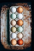 A dozen eggs of various colors are neatly arranged in a cardboard carton. The eggs vary in shades from white to brown, and green to blue, displaying a natural variety. The background consists of a rustic, textured surface with earthy tones.