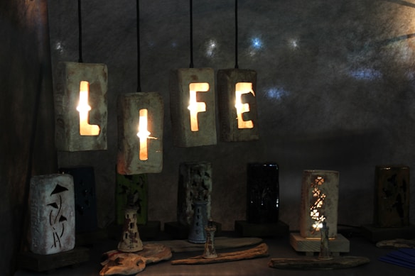 Illuminated block letters spelling out 'LIFE' are hanging against a textured dark background. Each letter is formed by hollow cuts in the blocks allowing light to shine through. Below, various artistic ceramic lamps sit on a surface, each with intricate cut-out designs and patterns that create an ambient glow.