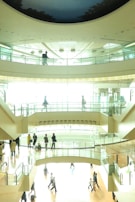 A modern multi-level indoor space with people walking and interacting on different floors. Glass barriers separate the levels, allowing light to fill the space, and the ceiling displays a circular design resembling a night sky. The scene is busy with individuals crossing paths, some carrying bags.