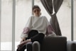 A person wearing decorative sunglasses and a large, loose-fitting sweater sits casually on a dark upholstered chair. The sweater is white with puffy pink sections on the sleeves. The setting appears to be indoors, with soft natural light filtering through large windows and grey curtains.