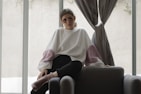 A person wearing decorative sunglasses and a large, loose-fitting sweater sits casually on a dark upholstered chair. The sweater is white with puffy pink sections on the sleeves. The setting appears to be indoors, with soft natural light filtering through large windows and grey curtains.