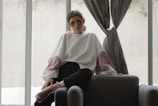A person wearing decorative sunglasses and a large, loose-fitting sweater sits casually on a dark upholstered chair. The sweater is white with puffy pink sections on the sleeves. The setting appears to be indoors, with soft natural light filtering through large windows and grey curtains.