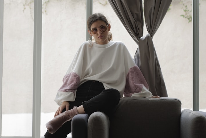 A person wearing decorative sunglasses and a large, loose-fitting sweater sits casually on a dark upholstered chair. The sweater is white with puffy pink sections on the sleeves. The setting appears to be indoors, with soft natural light filtering through large windows and grey curtains.