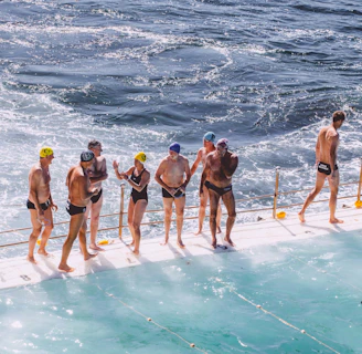 A group of people in swimwear are standing on the edge of a pool next to the ocean. Some are wearing swimming caps, and they appear to be preparing for or finishing a swim. The water in the pool and the ocean is choppy, and there is a railing separating the swimmers from the ocean.