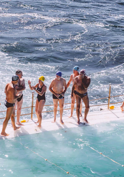 A group of people in swimwear are standing on the edge of a pool next to the ocean. Some are wearing swimming caps, and they appear to be preparing for or finishing a swim. The water in the pool and the ocean is choppy, and there is a railing separating the swimmers from the ocean.