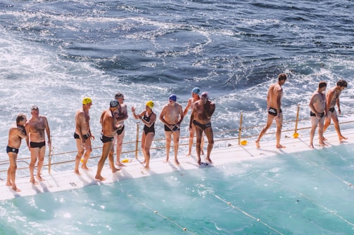 A group of people in swimwear are standing on the edge of a pool next to the ocean. Some are wearing swimming caps, and they appear to be preparing for or finishing a swim. The water in the pool and the ocean is choppy, and there is a railing separating the swimmers from the ocean.