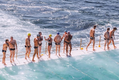 A group of people in swimwear are standing on the edge of a pool next to the ocean. Some are wearing swimming caps, and they appear to be preparing for or finishing a swim. The water in the pool and the ocean is choppy, and there is a railing separating the swimmers from the ocean.