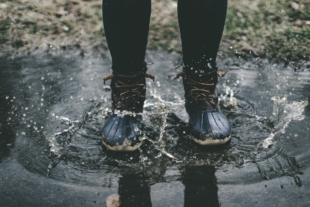 person wearing black duck boots stomping on body of water