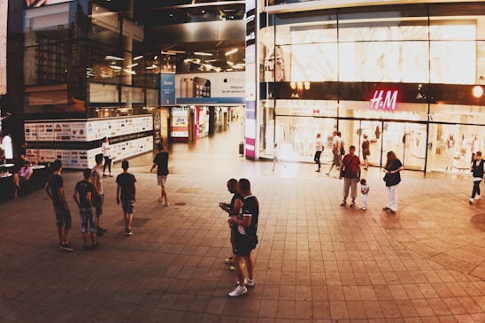 People walk and gather around a brightly lit shopping mall entrance. The storefronts display various brand signs, including a prominent H&M store. The scene appears lively with individuals engaged in activities such as walking, standing, and using mobile phones.