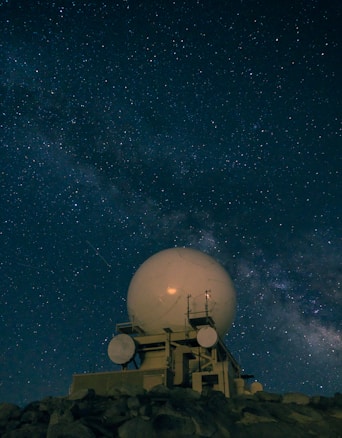 A large, spherical radar antenna sits atop a rocky hill under a vast, starry night sky. The structure includes supporting equipment and is silhouetted against the dense Milky Way galaxy visible above.
