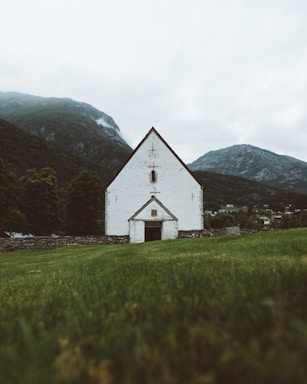 A white church with a dark roof stands in the foreground of a serene, green landscape. The church is positioned centrally, facing forward, with a stone wall surrounding it. Behind the church are tall, forested mountains partially covered in mist or low clouds. The sky above is overcast, contributing to a subdued and calm atmosphere.