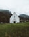 A white church with a dark roof stands in the foreground of a serene, green landscape. The church is positioned centrally, facing forward, with a stone wall surrounding it. Behind the church are tall, forested mountains partially covered in mist or low clouds. The sky above is overcast, contributing to a subdued and calm atmosphere.