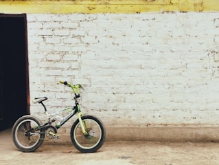 A BMX bicycle with a black and green frame is parked against a white brick wall. The ground is covered in dirt and the wall is topped with a yellow stripe. There is an open door to the left leading into darkness.