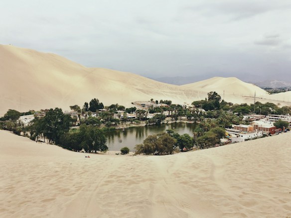 A picturesque oasis surrounded by vast sand dunes with a small body of water in the center. Lush greenery and a collection of buildings, likely a village or small settlement, surround the water. The landscape is expansive with rolling dunes under a cloudy sky.