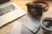 A still life scene on a wooden desk features a vintage camera, a laptop, photographs, and a steaming cup of coffee. The camera and photographs evoke a sense of nostalgia or travel.