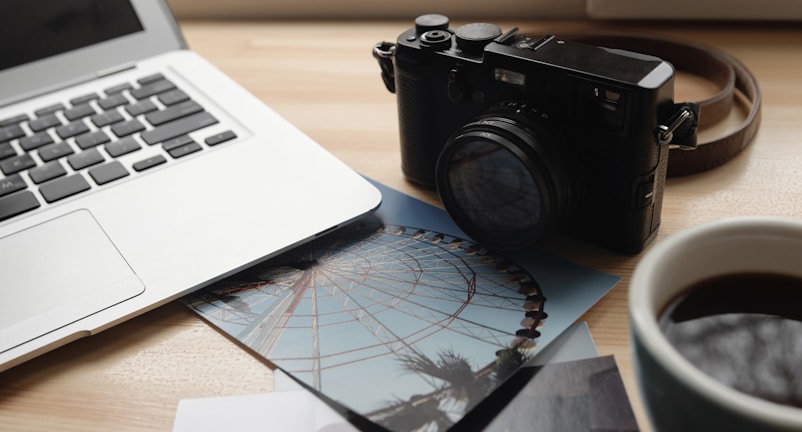 A tabletop scene featuring an open laptop computer, a black vintage-style camera, printed photographs, and a cup of coffee. The photographs show a ferris wheel against a blue sky. The setting conveys a mix of technology, photography, and relaxation.