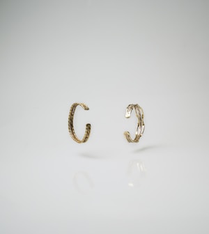 Two gold-toned, textured open rings or bracelets positioned vertically against a smooth, light background, with subtle reflections visible on the surface beneath.