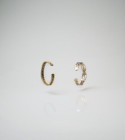 Two gold-toned, textured open rings or bracelets positioned vertically against a smooth, light background, with subtle reflections visible on the surface beneath.