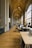 The image depicts a spacious, ornately decorated workspace with high ceilings and arched windows. The ceiling features intricate detailing and several large, elegant chandeliers. The floors are made of polished wood, and several desks are arranged along the corridor, equipped with computers and office chairs. Natural light floods the space, adding warmth and brightness.