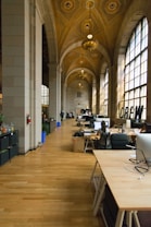 The image depicts a spacious, ornately decorated workspace with high ceilings and arched windows. The ceiling features intricate detailing and several large, elegant chandeliers. The floors are made of polished wood, and several desks are arranged along the corridor, equipped with computers and office chairs. Natural light floods the space, adding warmth and brightness.