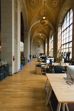 The image depicts a spacious, ornately decorated workspace with high ceilings and arched windows. The ceiling features intricate detailing and several large, elegant chandeliers. The floors are made of polished wood, and several desks are arranged along the corridor, equipped with computers and office chairs. Natural light floods the space, adding warmth and brightness.