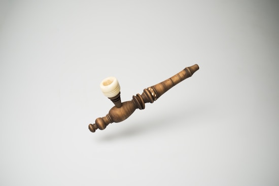 A handcrafted wooden pipe with intricate carvings and a cream-colored bowl is placed against a plain, light background. The pipe appears ornate and polished, showcasing the craftsmanship involved in its creation.