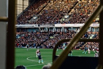 A large crowd fills an outdoor stadium, intently watching a soccer match. Players in uniforms are actively engaging in the game on the field, while the sun casts shadows over the scene. The stands are packed with spectators, with advertisements visible on the walls behind them. The image is framed by railings, suggesting it's taken from a higher vantage point within the structure.