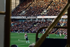 A large crowd fills an outdoor stadium, intently watching a soccer match. Players in uniforms are actively engaging in the game on the field, while the sun casts shadows over the scene. The stands are packed with spectators, with advertisements visible on the walls behind them. The image is framed by railings, suggesting it's taken from a higher vantage point within the structure.