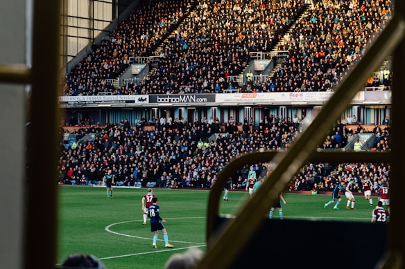 A large crowd fills an outdoor stadium, intently watching a soccer match. Players in uniforms are actively engaging in the game on the field, while the sun casts shadows over the scene. The stands are packed with spectators, with advertisements visible on the walls behind them. The image is framed by railings, suggesting it's taken from a higher vantage point within the structure.