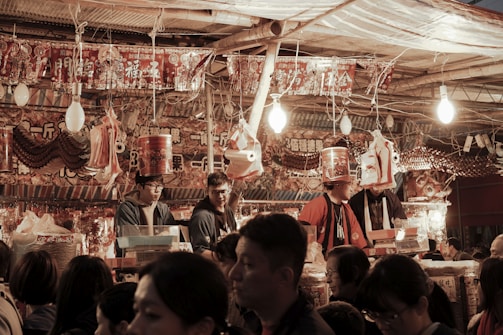A bustling market stall is crowded with people, with vibrant red and orange decorations featuring Chinese characters hanging above them. The scene is warmly lit by hanging bulbs, and several vendors engage with customers. Various packaged goods and traditional items are displayed around the stall.