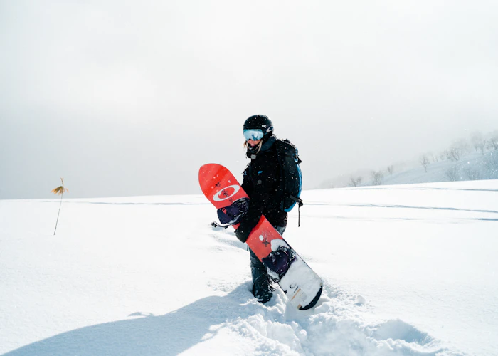 Rider in black gear carrying a red snowboard across fresh snow