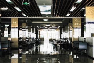 A spacious, modern cafeteria with rows of tables and chairs neatly arranged in a symmetrical pattern. The ceiling features a grid of rectangular lights and wooden beams, and there are reflective tiles on the floor adding to the symmetrical aesthetic. Natural light floods in from large windows at the end of the room, creating a bright and open atmosphere.