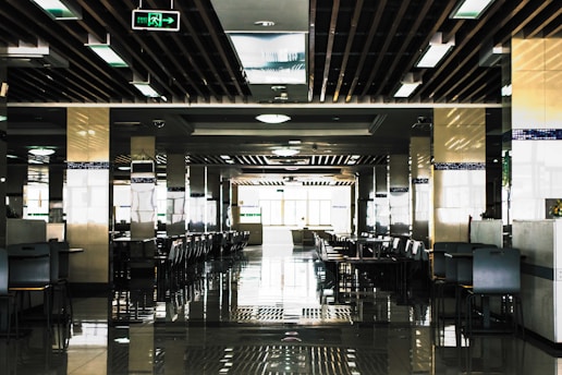 A spacious, modern cafeteria with rows of tables and chairs neatly arranged in a symmetrical pattern. The ceiling features a grid of rectangular lights and wooden beams, and there are reflective tiles on the floor adding to the symmetrical aesthetic. Natural light floods in from large windows at the end of the room, creating a bright and open atmosphere.
