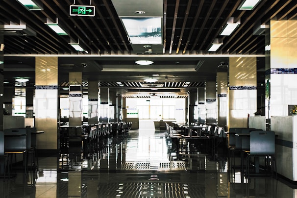 A spacious, modern cafeteria with rows of tables and chairs neatly arranged in a symmetrical pattern. The ceiling features a grid of rectangular lights and wooden beams, and there are reflective tiles on the floor adding to the symmetrical aesthetic. Natural light floods in from large windows at the end of the room, creating a bright and open atmosphere.